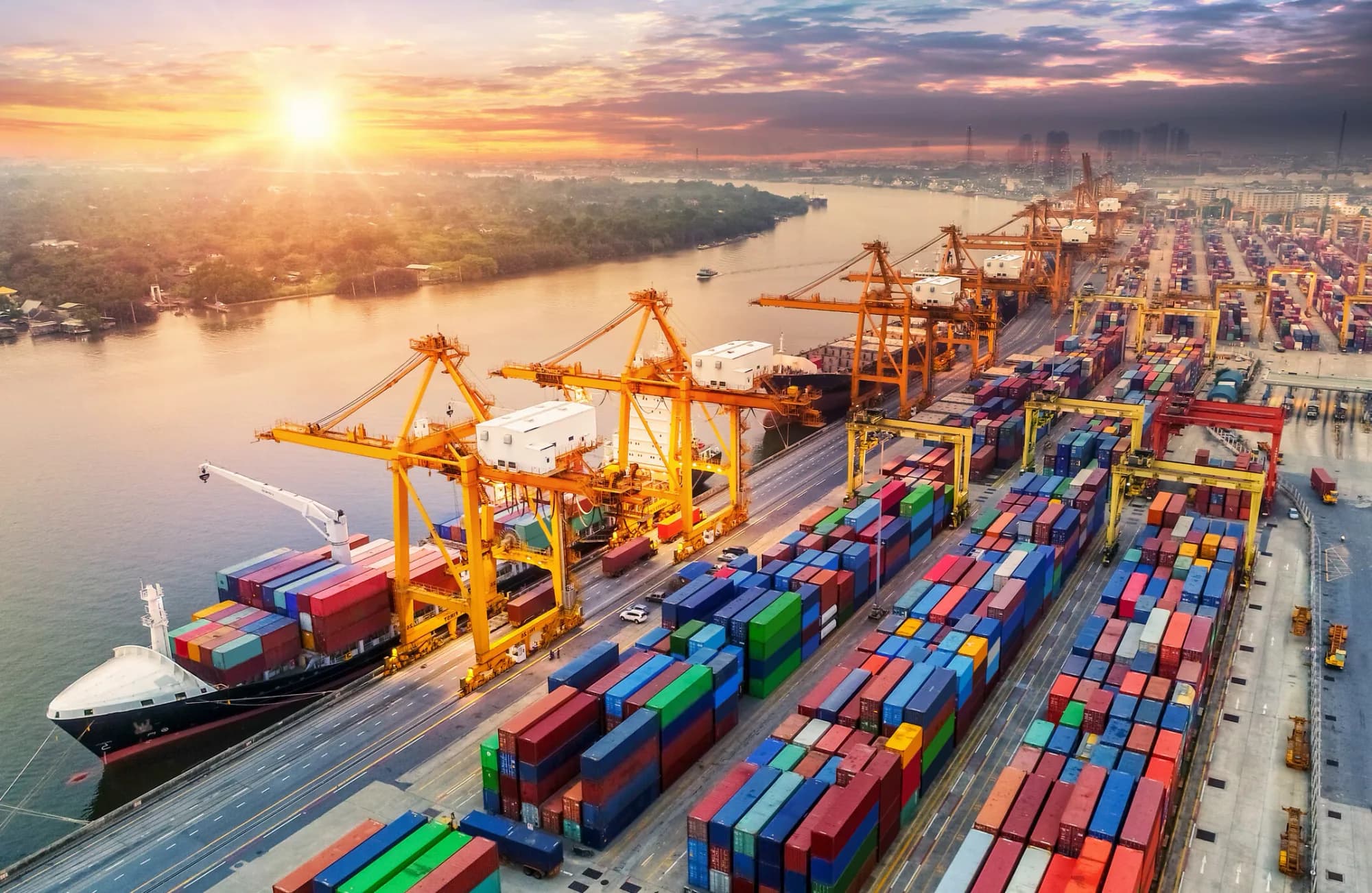 An aerial view of a container port at sunset, with gantry cranes working a container ship and rows of stacked containers along the waterfront.