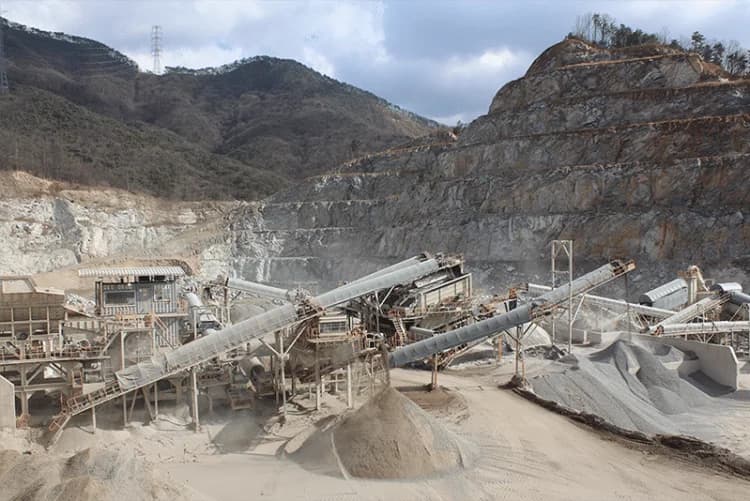 A rock quarry with a crushing and screening plant, stacker conveyors, and aggregate stockpiles beneath a terraced rock face.