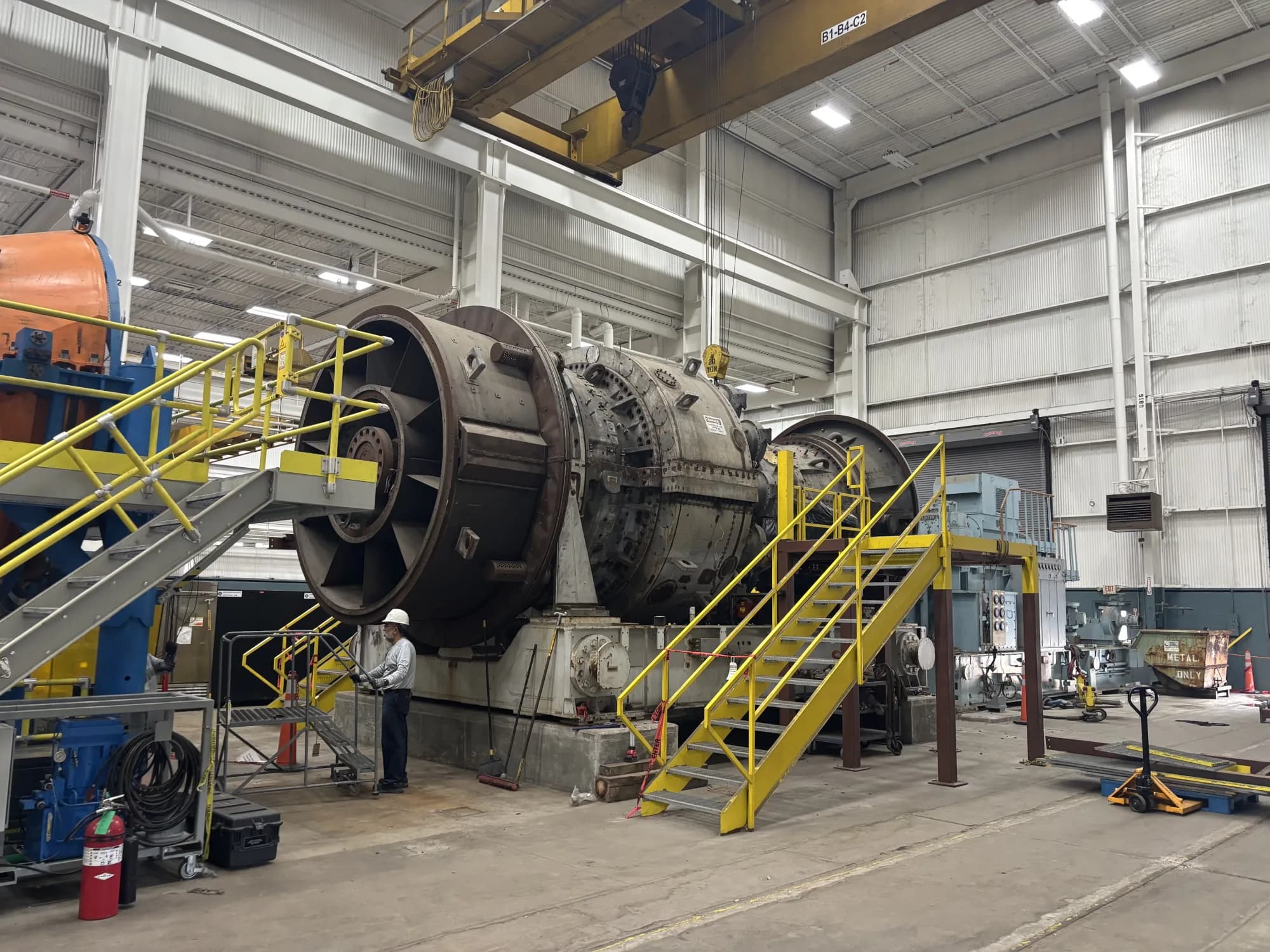 A large industrial rotor and casing assembly under overhaul in a high-bay machine shop, with an overhead bridge crane, yellow access stairs, and a technician standing at its base for scale.