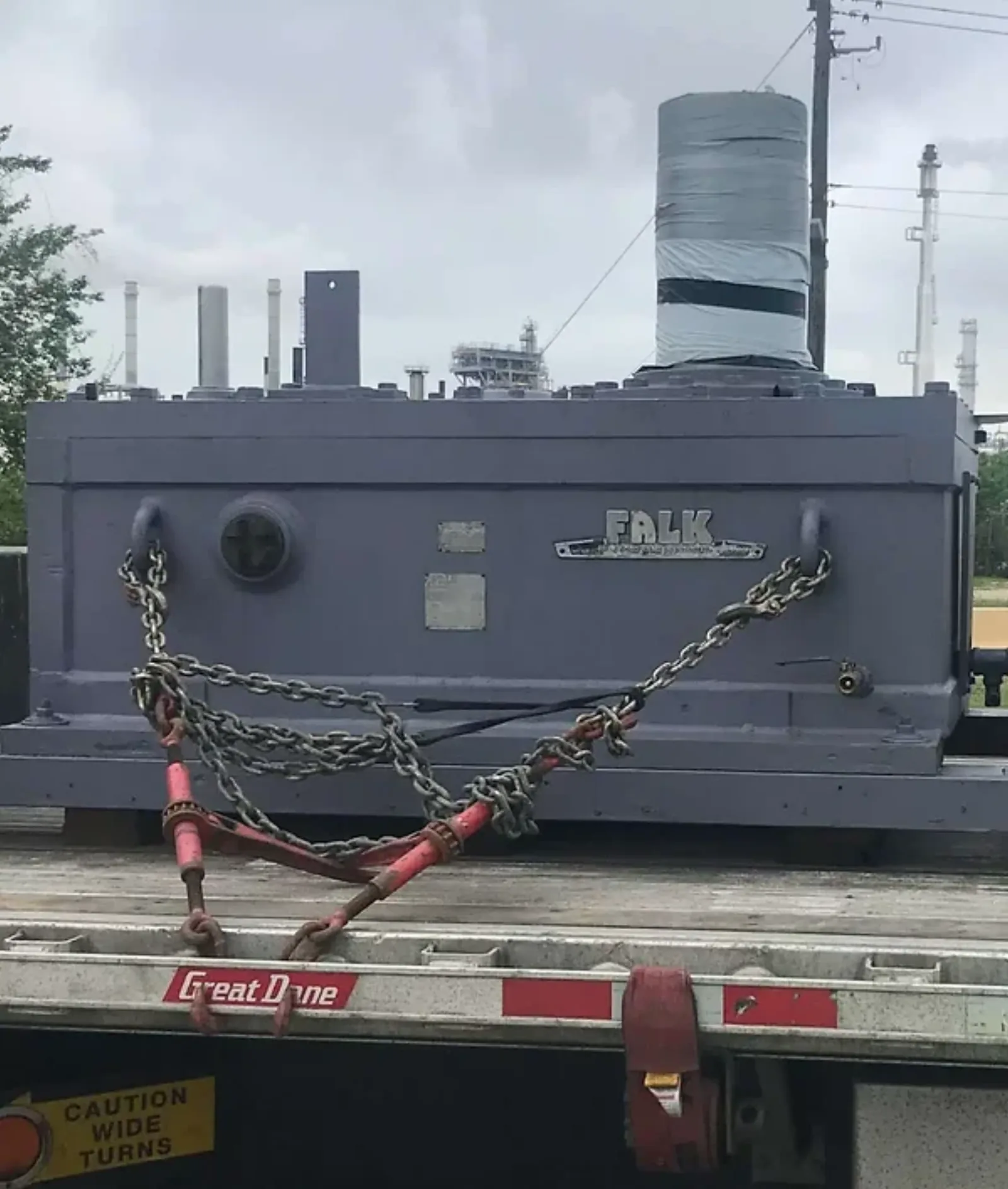 A rebuilt Falk industrial gearbox chained to a flatbed trailer with a petrochemical plant in the background.