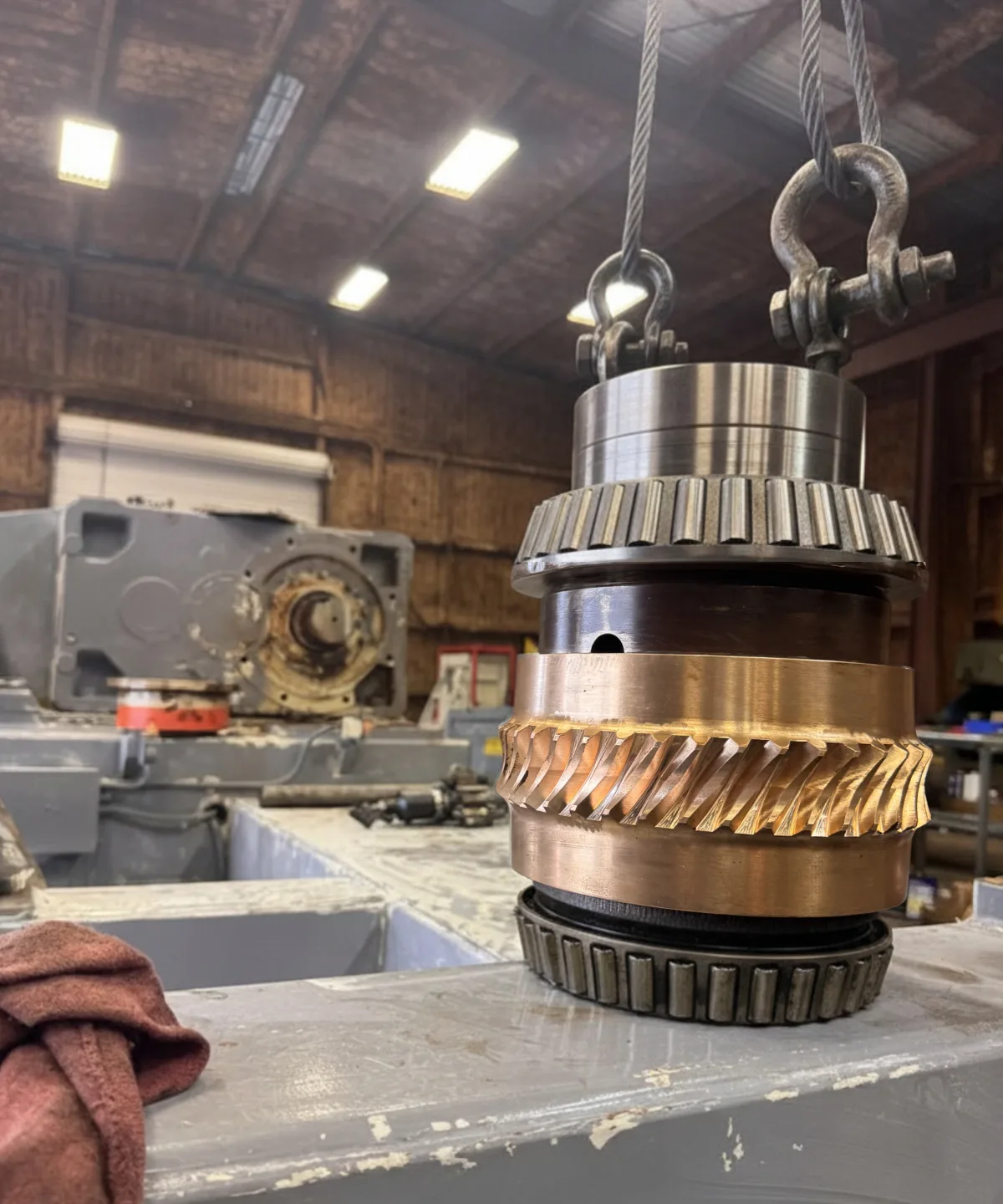 A bronze bevel gear and bearing stack rigged and hanging by chain, with a worn gearbox housing in the background.
